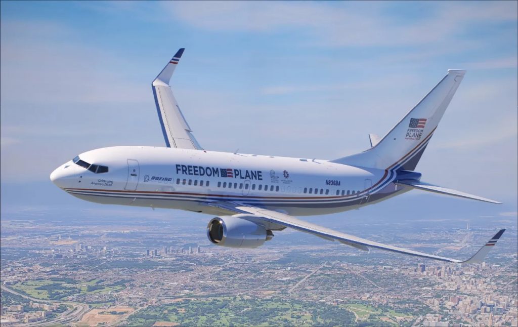 A photo of a white passenger airplane labeled “Freedom Plane” flying above a sprawling city landscape, with red and blue stripes along the fuselage and the Boeing logo near the front.