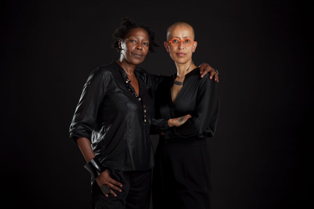 two women stand together, each wearing black, they are in a partial embrace, with the woman on the left holding her arm around the other woman's shoulder. they stare directly at the camera and the background behind is black