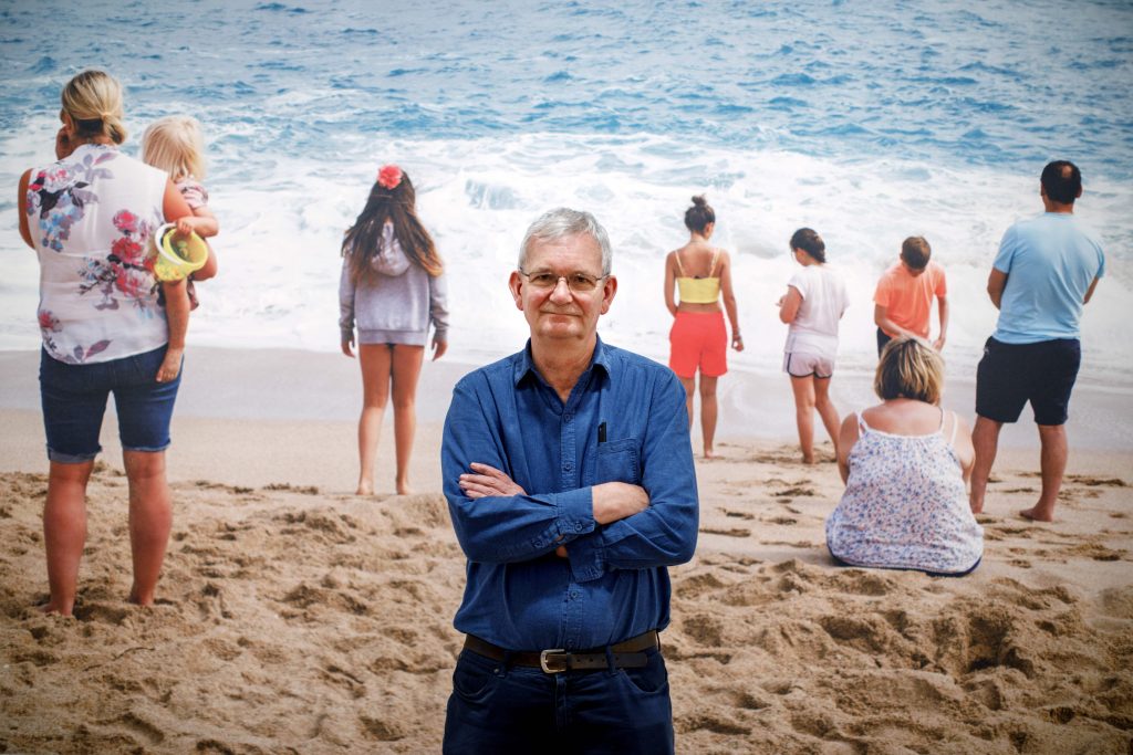 Man standing with arms crossed on a sandy beach while a group of people face the shoreline waves in the background.