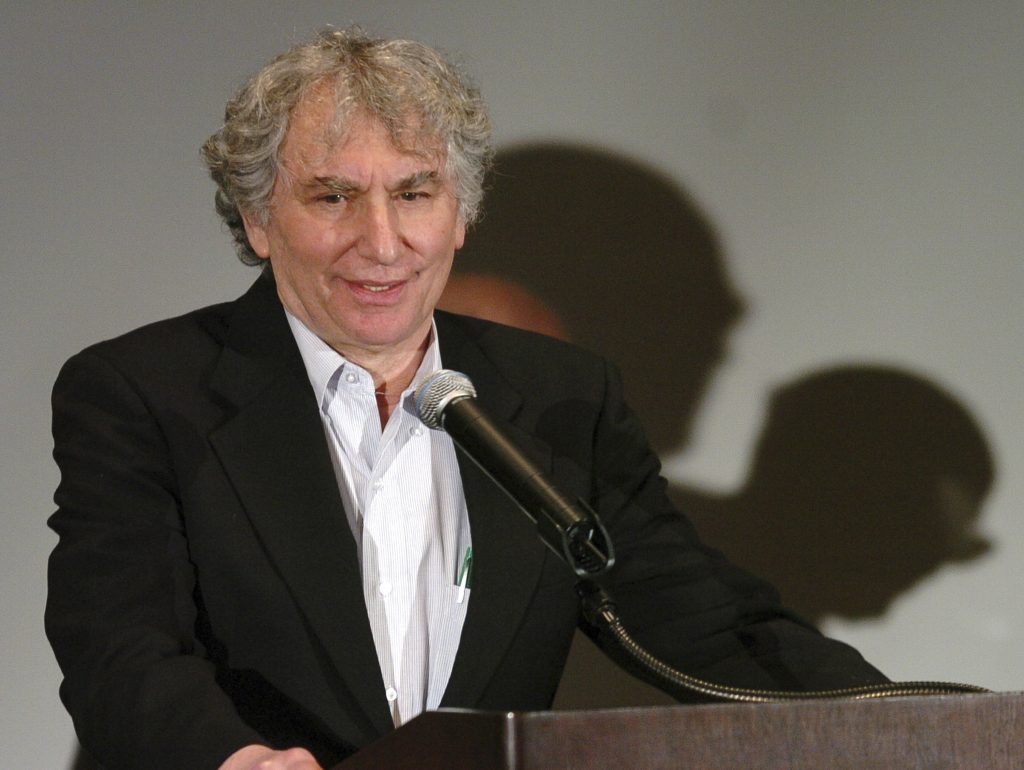 Older man with curly gray hair speaking at a podium with a microphone, wearing a dark blazer and light shirt.