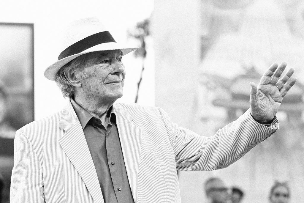 Black-and-white photograph of an older man in a striped blazer and panama hat raising his hand in greeting during a public cultural event.