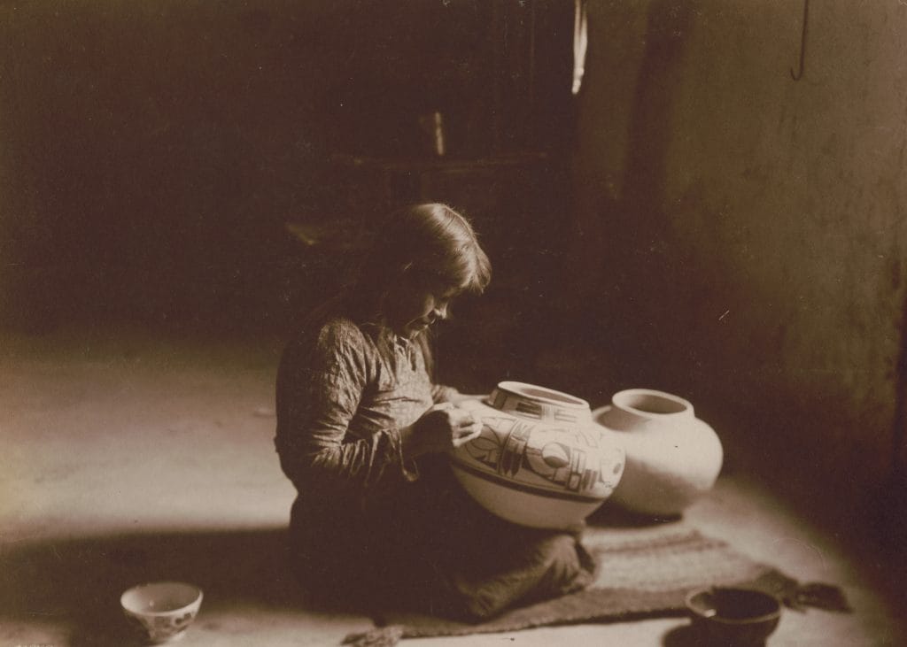 An older Native American woman sits surrounded by finished ceramic vessels—bowls, jars, and seed pots—arranged on a woven textile. She holds a decorated plate featuring bold, geometric motifs typical of Pueblo pottery. The designs—stylized birds, feathers, and abstract forms in black-on-white—are characteristic of communities such as Acoma, Hopi, or Laguna Pueblo. The setting and presentation suggest both craftsmanship and cultural continuity, emphasizing the artist’s role as keeper of tradition.