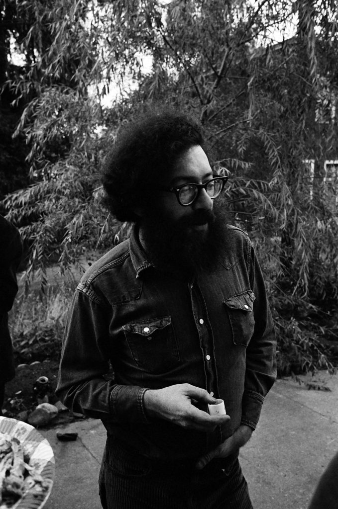 Black-and-white candid portrait of a man with glasses and a full beard standing outdoors in a natural setting, wearing a denim shirt and holding a small object, evoking 1970s counterculture style.