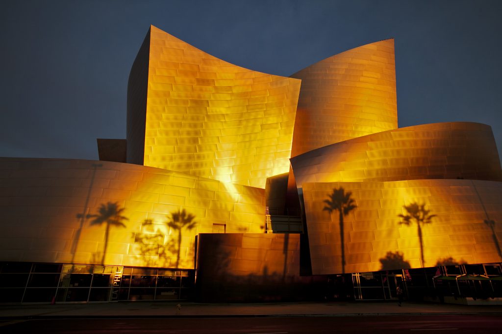 The Walt Disney Concert Hall glows golden at sunset, with palm tree shadows stretching across its curving metal facade.