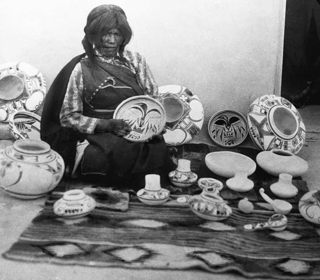 An older Native American woman sits surrounded by finished ceramic vessels—bowls, jars, and seed pots—arranged on a woven textile. She holds a decorated plate featuring bold, geometric motifs typical of Pueblo pottery. The designs—stylized birds, feathers, and abstract forms in black-on-white—are characteristic of communities such as Acoma, Hopi, or Laguna Pueblo. The setting and presentation suggest both craftsmanship and cultural continuity, emphasizing the artist’s role as keeper of tradition.