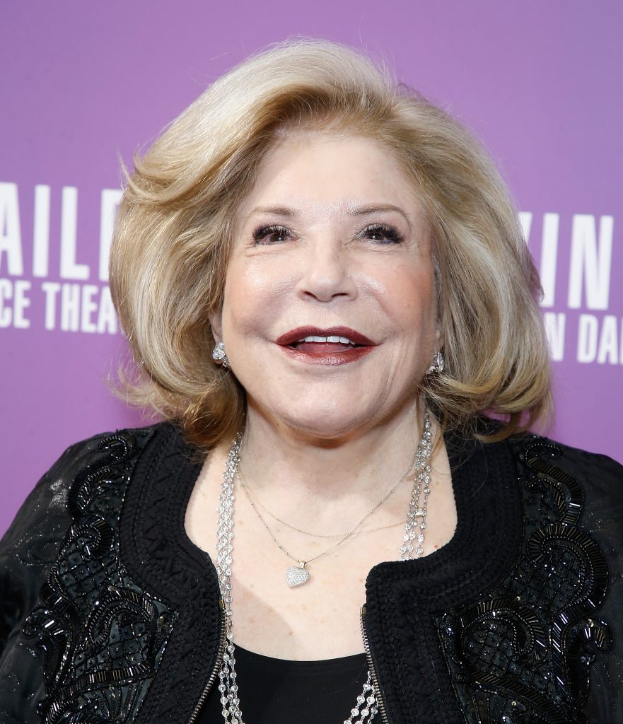Smiling older woman with styled blonde hair posing against a purple backdrop, wearing a black embroidered jacket and layered silver necklaces.