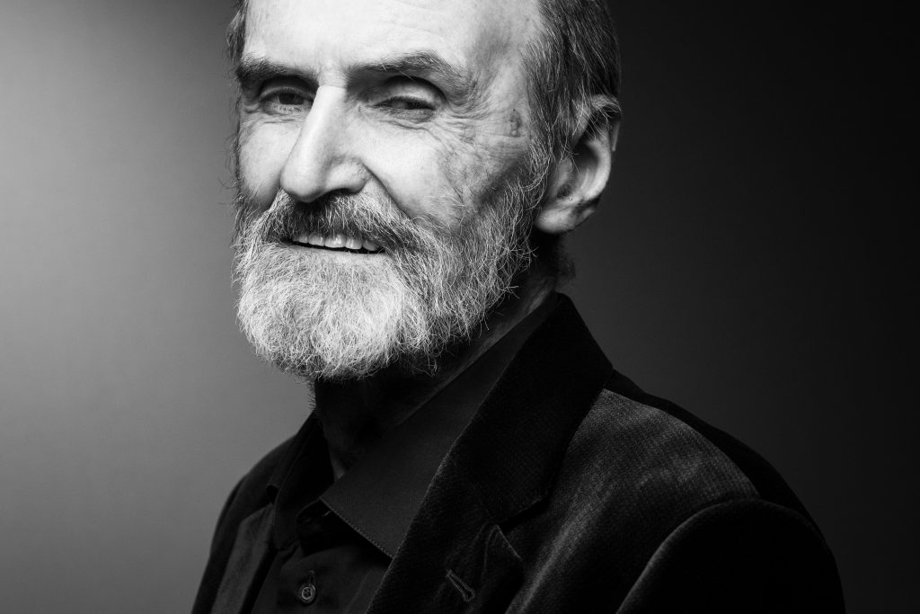 Black-and-white portrait of an older man with a full beard and slight smile, wearing a dark collared shirt and jacket against a softly lit studio background.