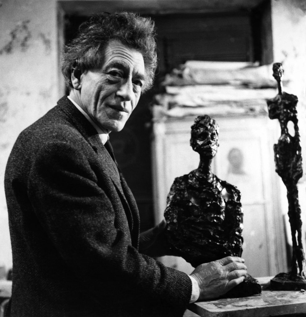 Black-and-white portrait of Giacometti holding textured bronze figure in studio, expressive face, artworks visible nearby