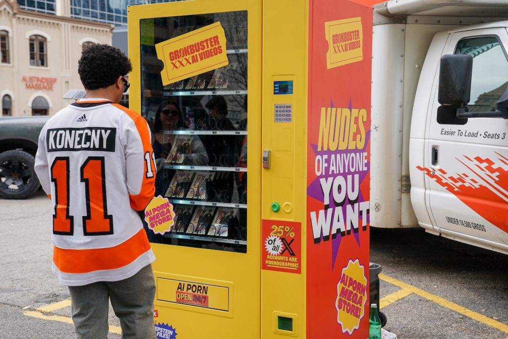 A person in a hockey jersey stands before a yellow vending machine labeled "Grokbuster XXX AI Videos," featuring provocative advertisements for AI-generated adult content.