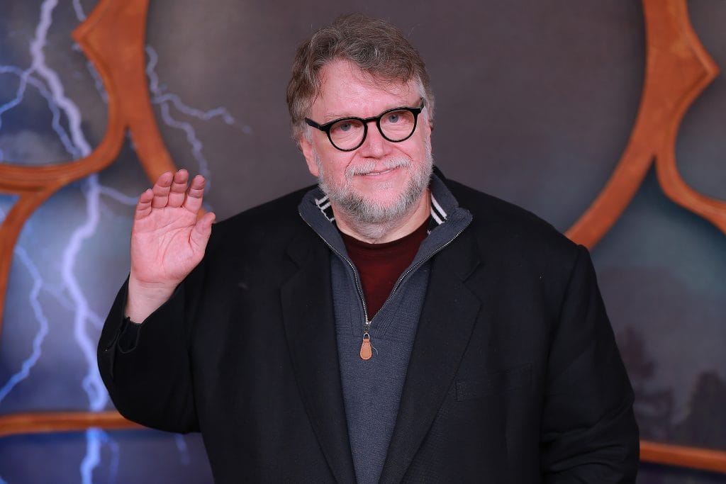 A photograph of Guillermo del Toro waving and smiling in a black cardigan before a red and blue movie poster