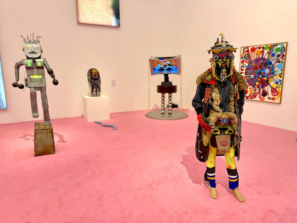 Multiple eclectic, humanoid sculptures and a video installation are spread across a pink carpeted gallery, featuring a variety of materials, vibrant patterns, and mechanical elements.