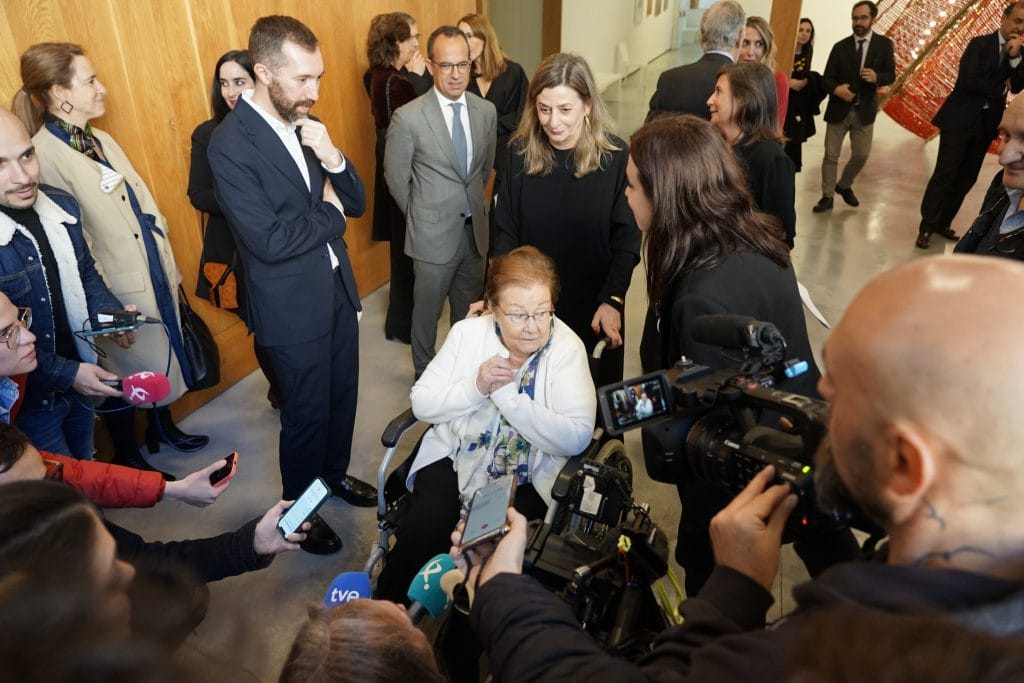 A photo of an older white woman with short reddish hair sitting in a wheelchair at a public event, surrounded by journalists holding microphones, cameras, and smartphones toward her. She wears a white cardigan over a floral blouse and appears to be responding to the crowd. Around her stand several well-dressed attendees and officials in a modern gallery space.