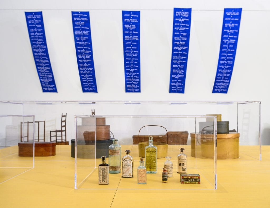 an image of the artwork installed hanging from the ceiling above several other Shaker artifacts enclosed in a glass vitrine