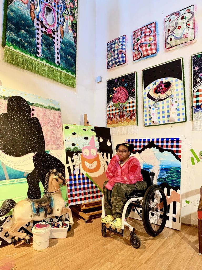 a black woman wearing a red sweater and green pants sits in a wheelchair surrounded by colorful textured artworks