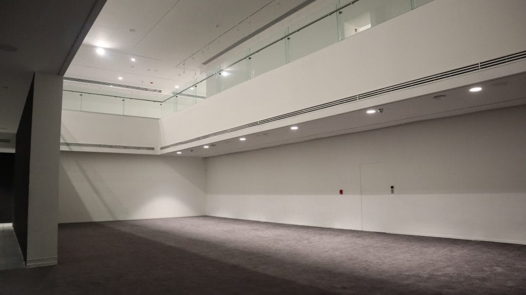 A sparse, white-walled gallery interior with high ceilings, recessed lighting, and a mauve carpet covering the floor, forming a minimal exhibition space.