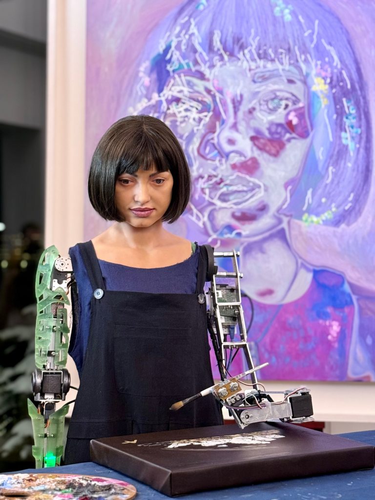 a humanoid robot in a bob and overalls stands with her robotic arm holding a paintbrush, behind her is a purple artwork image of her, she is standing in a public interior space