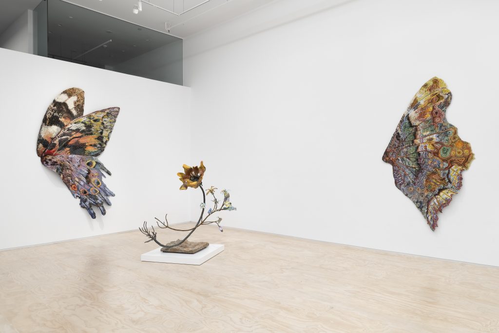 Installation view of a white-walled gallery with two large, richly patterned butterfly-shaped textile wall works—one spread across the left wall and another on the right—framing a small sculptural piece on a low white platform in the center, resembling a branching stem with flowers.