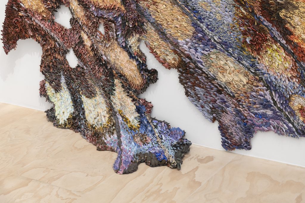 Close-up of a large, layered textile wall installation by Rebecca Manson composed of thousands of small, petal-like fabric pieces in earthy reds, browns, blues, and purples, forming an abstract, winglike shape that extends down to the gallery floor.