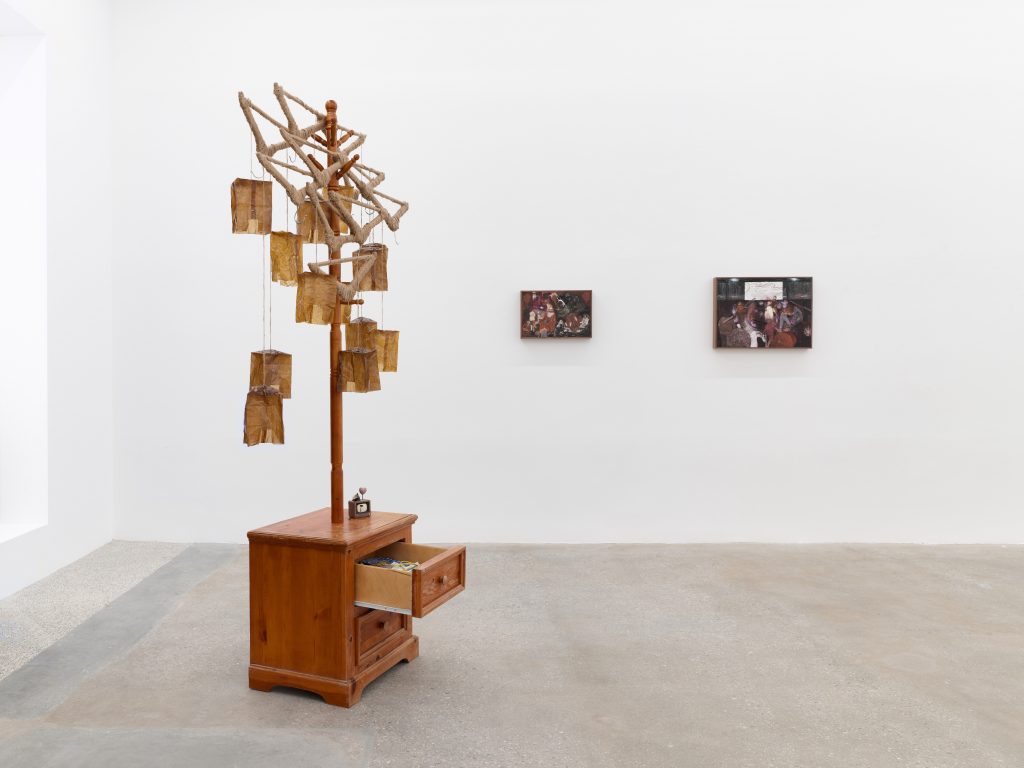 "Veronica Fernandez: Prey" gallery installation view featuring a sculptural work made from a wooden cabinet with drawers and a vertical post supporting a rope lattice structure with hanging brown paper elements. Two small framed paintings hang on the white wall behind it in a minimalist gallery space with a concrete floor.