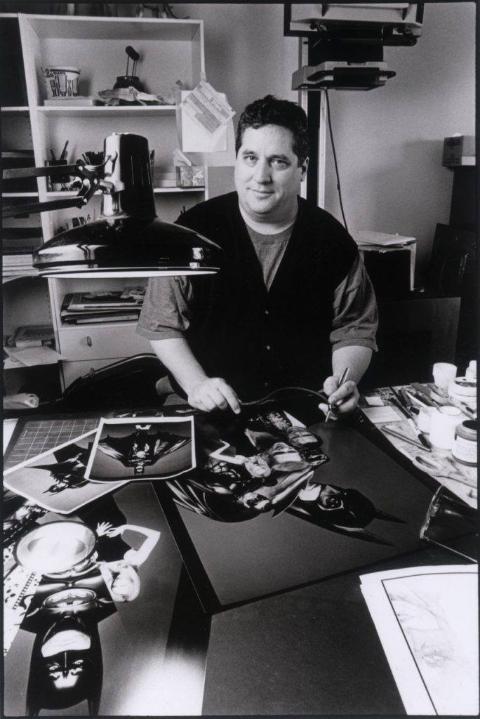Black-and-white photograph shows artist John Alvin at drafting table, painting Batman poster artwork in studio.