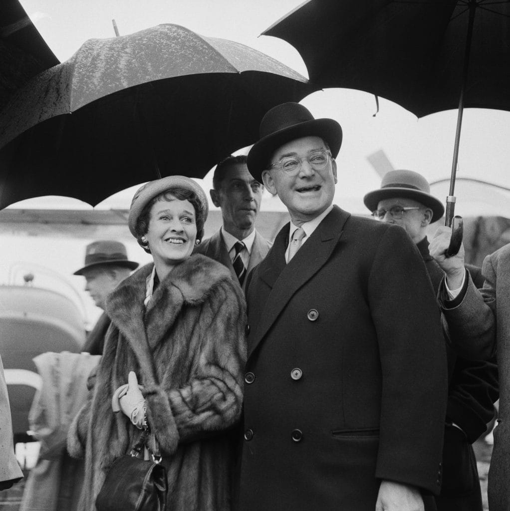 Well-dressed couple, John Hay Whitney and Betsey Cushing Whitney, under umbrellas in rain, smiling, surrounded by men in suits during public event