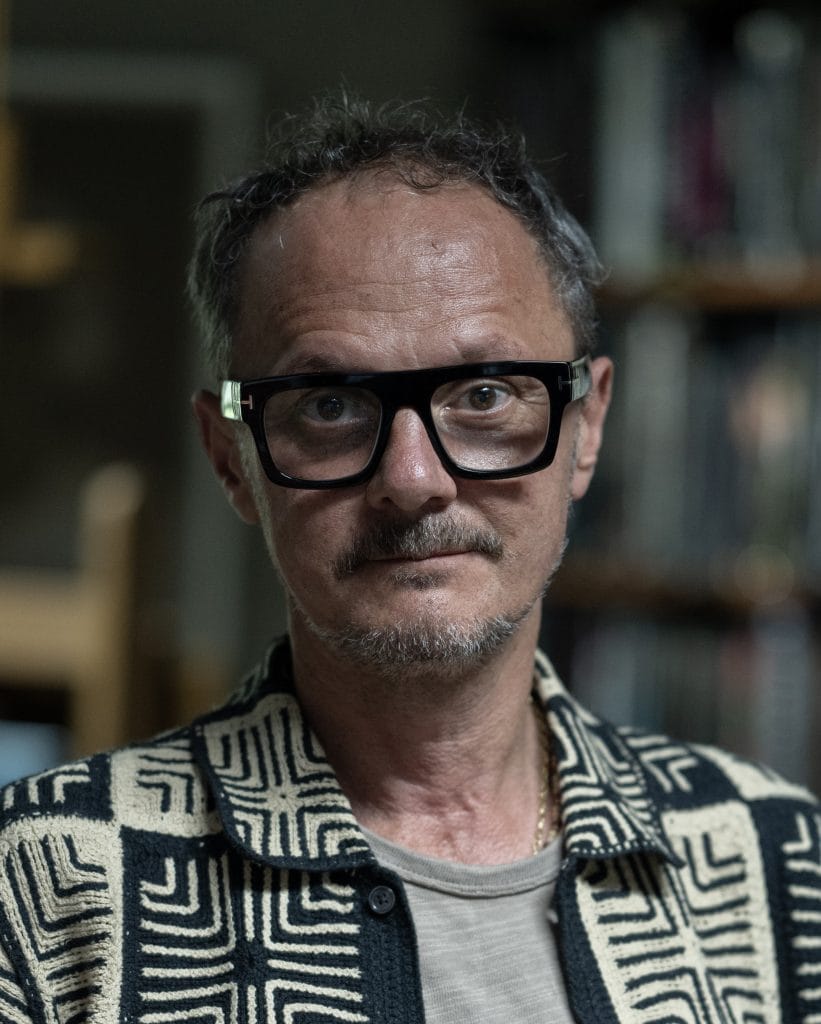 Portrait of artist Jonathan Yeo wearing glasses and patterned jacket before blurred studio bookshelves.