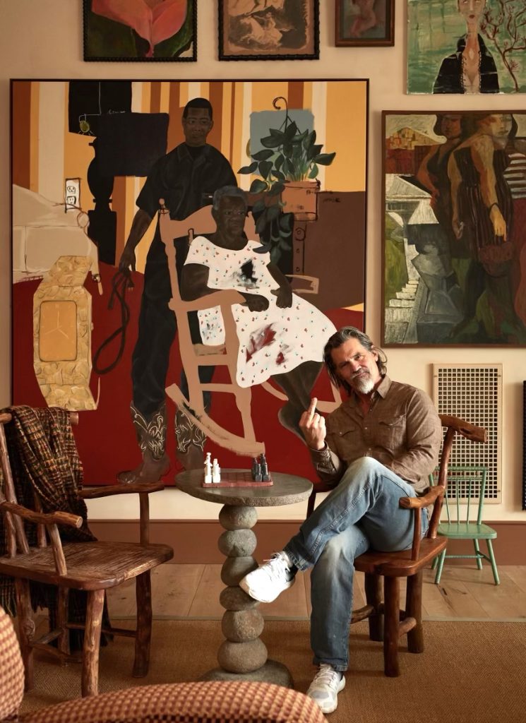 A photo of a white middle-aged man with gray hair and a gray beard sitting cross-legged in a wooden chair, wearing a brown button-down shirt, blue jeans and white sneakers, raising his middle finger toward the camera. He is seated at a small round stone table with a chessboard on top, in front of a large colorful painting depicting a Black man standing behind an older Black woman seated in a rocking chair, surrounded by multiple framed artworks on the wall.