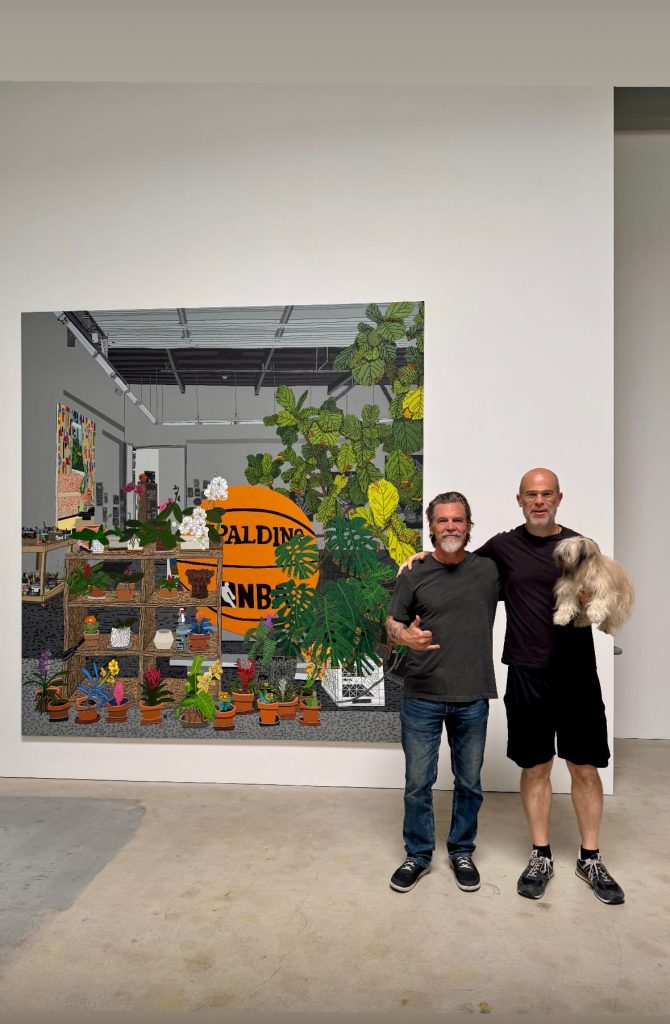 A photo of two white middle-aged men standing arm in arm in a gallery space in front of a large colorful painting filled with potted plants and a giant orange basketball. One man has gray hair and a gray beard and wears a dark T-shirt and jeans, making a shaka hand gesture, while the other is bald, wearing glasses, a dark T-shirt and black shorts, holding a small fluffy tan dog.