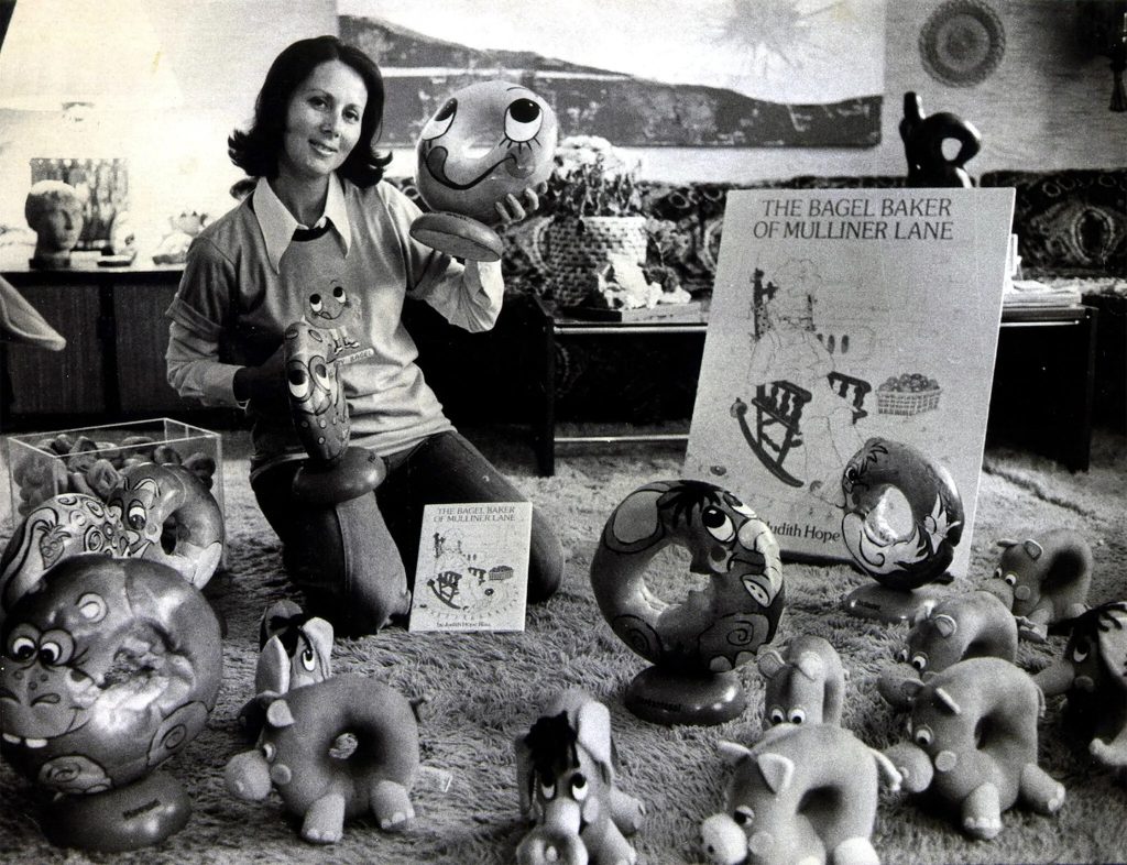Black-and-white photo of a woman kneeling on a carpet, holding whimsical bagel-shaped characters surrounded by illustrated sculptures and a display for "The Bagel Baker of Mulliner Lane."