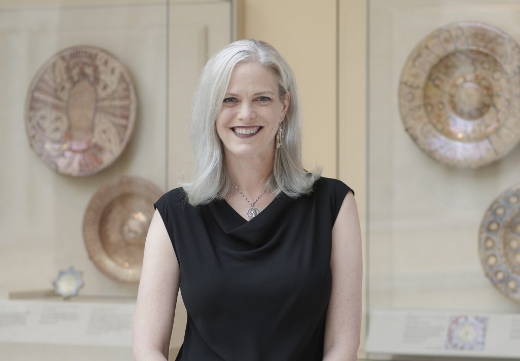 Woman standing in a museum gallery with historic ceramic plates displayed behind her, smiling in a contemporary portrait highlighting art and cultural heritage.