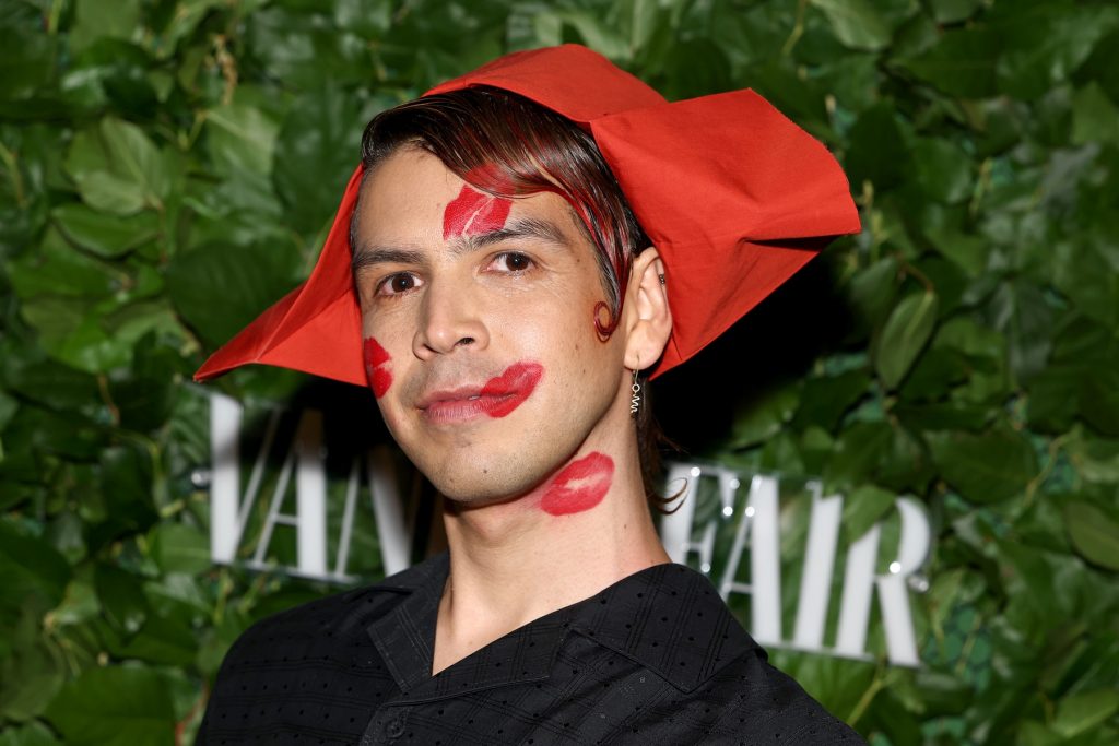 Close-up of a person with red lipstick kiss marks on their face and neck, wearing a structured red fabric headpiece and a black shirt.