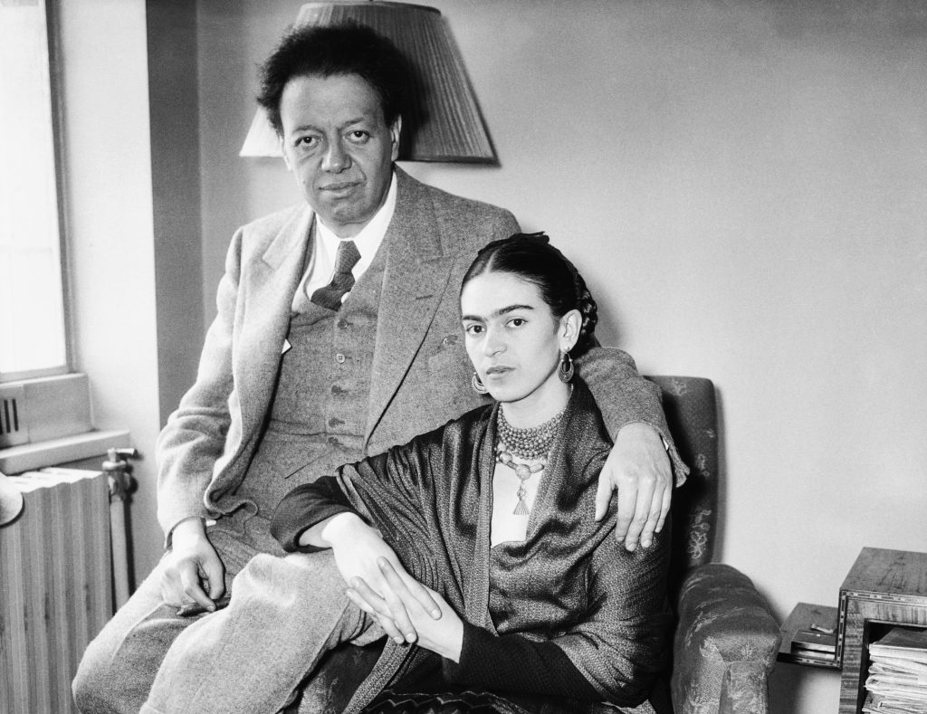 Frida Kahlo seated beside Diego Rivera, his arm around her, in intimate black-and-white portrait.