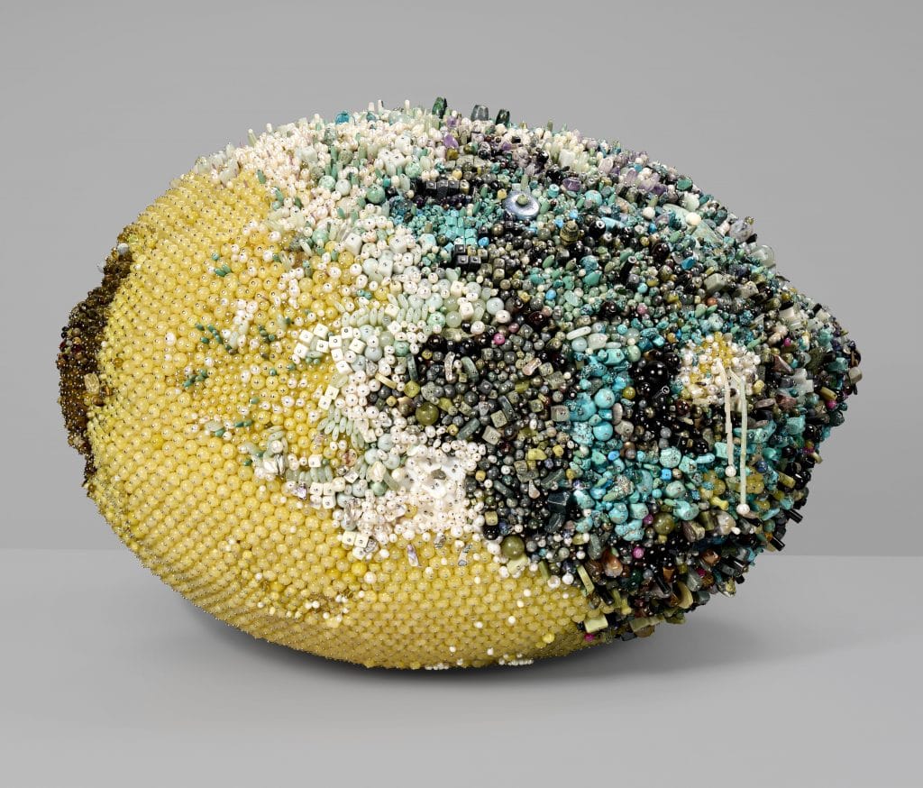 Beaded sculptural object resembling a lemon, covered in dense clusters of multicolored stones and pearls against a neutral background.