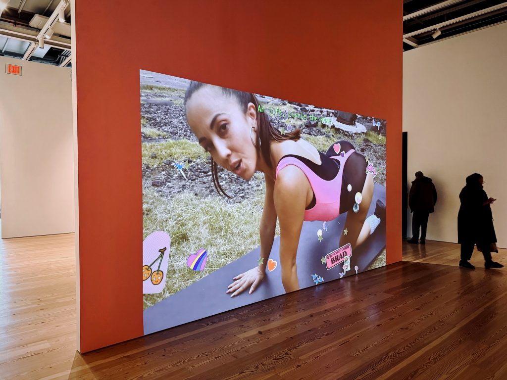 A video of a woman in pink athletic gear performing a workout is projected onto a large salmon-colored wall in a bright, modern art gallery.
