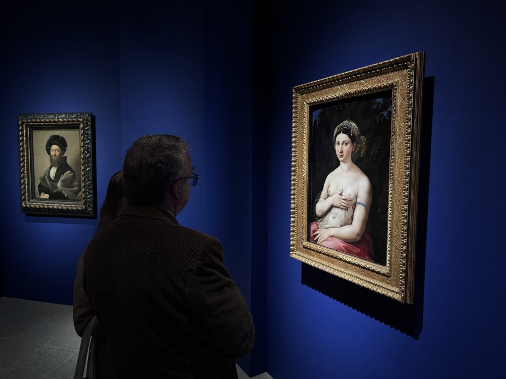 A man views Raphael’s La Fornarina in a museum. The portrait of a semi-nude woman hangs on a deep blue wall beside another framed painting.