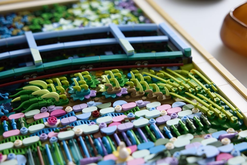 A photo of a close-up detail of a LEGO mosaic artwork showing a green arched bridge above a densely textured pond, with layered LEGO pieces forming lily pads, stems, flowers, and reeds in shades of green, blue, pink, and lavender, emphasizing the intricate construction and surface depth.