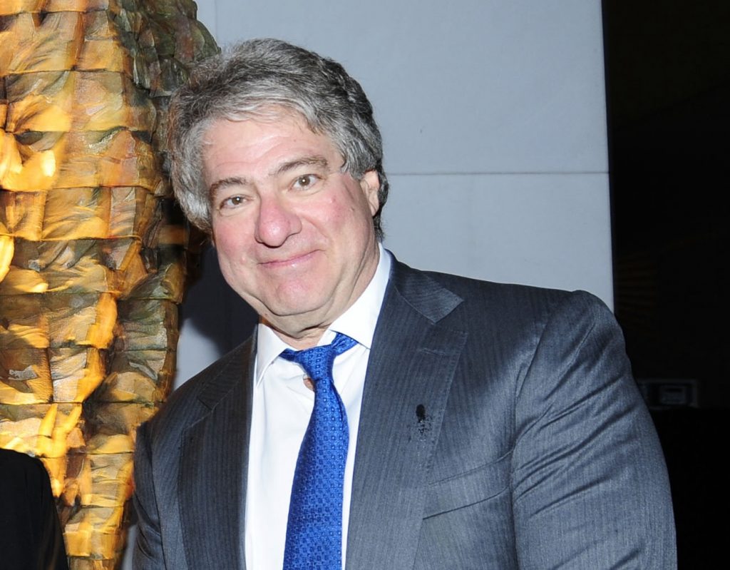 A photo of a middle-aged white man with thick gray hair wearing a gray suit and bright blue tie, smiling while standing indoors near a textured wall sculpture.