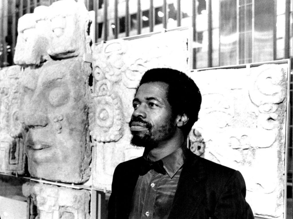 A black and white photo of a young Black man with a short afro and trimmed beard standing in front of large carved stone reliefs. He wears a collared shirt and jacket and gazes slightly upward with a thoughtful expression.