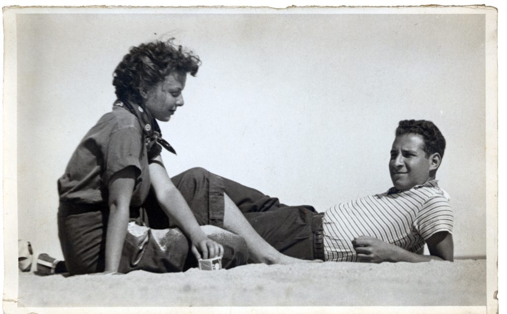 Black-and-white photograph of a young woman and man relaxing on a sandy beach, facing each other; the woman sits upright while the man reclines, both dressed casually against a clear sky.