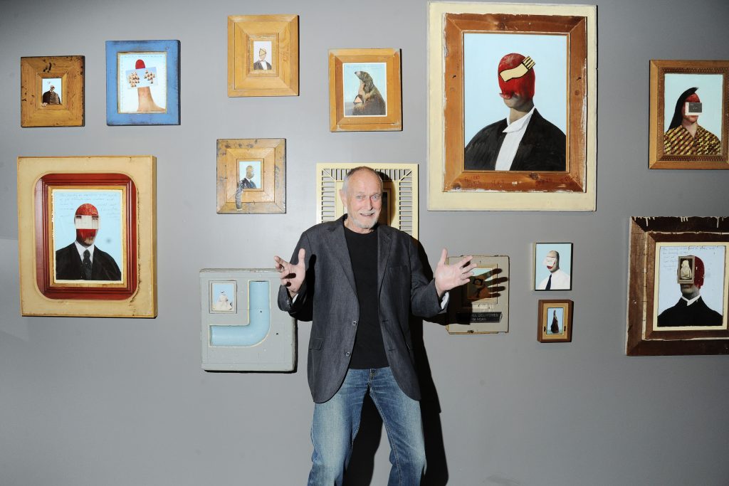 A photo of an older white man with short gray hair and a beard standing enthusiastically in front of a wall filled with framed surreal portrait paintings. He wears a dark blazer, black shirt, and jeans, gesturing broadly with both hands in a lively gallery setting.