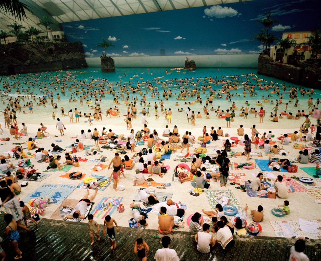 a large crowd of people appear to be at the beach but we can tell that in fact it is a fake beach inside a large building