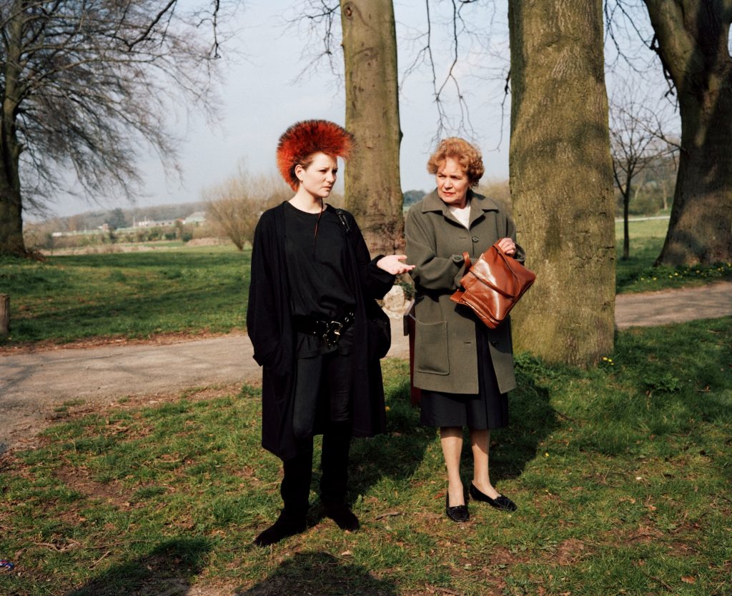 two women stand outside in a grassy park, one is a young punk with a dyed red mohawk and the other is a more prim older woman, they are an amusing contrast