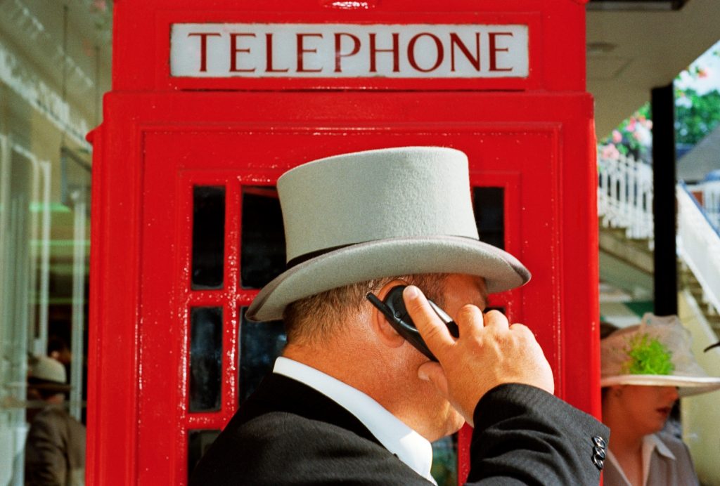 a man in a grey top hat holds a phone up to his ear, behind him is a shiny red telephone box with 'Telephone' written on it