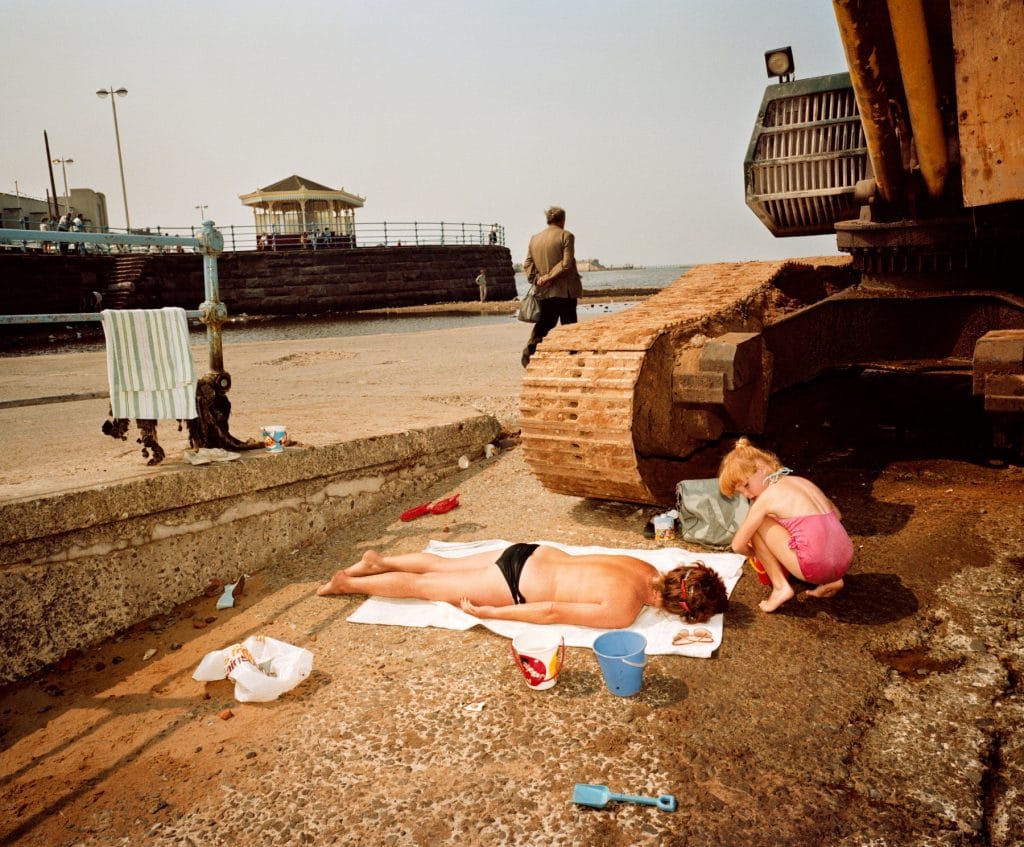 a beach scene in which a topless sunbathing woman lies face down on a towel and a child plays near her, they are next to a large tractor, creating a comical pairing