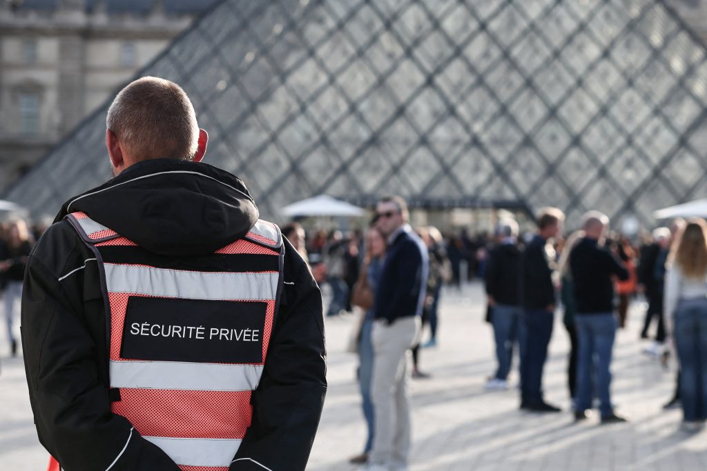 Private security guard in reflective vest watches over crowd outside Louvre Pyramid entrance in bright daylight.