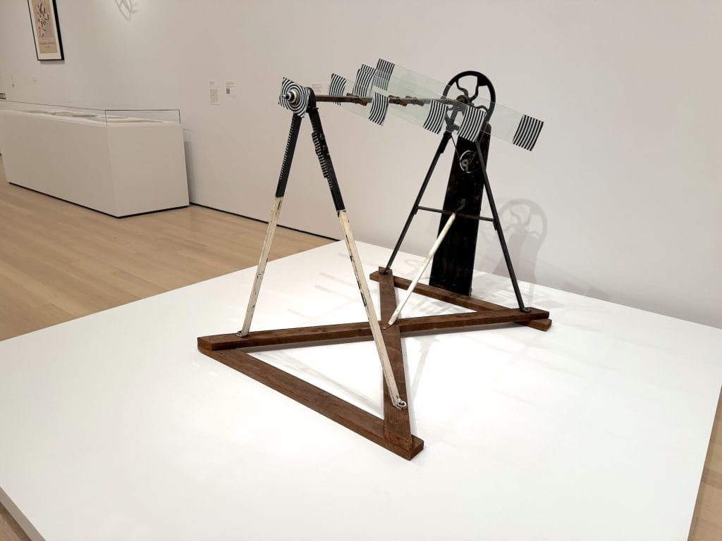 A motorized kinetic sculpture featuring five glass plates with black-and-white stripes, mounted on a metal axle and wooden frame in a gallery setting.
