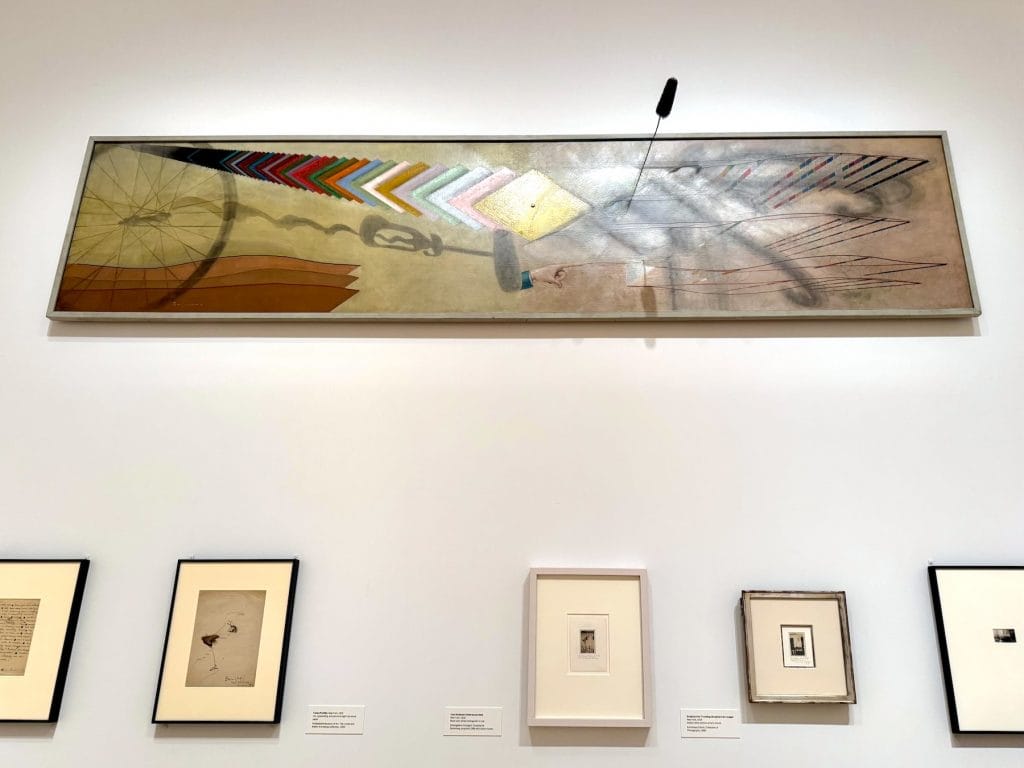 Large horizontal oil painting Tu m' by Marcel Duchamp, featuring various shadows and colorful receding shapes, mounted above a row of smaller framed works.
