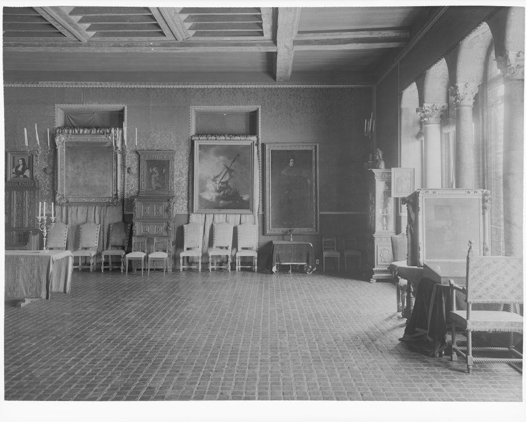 Black-and-white photograph of an ornate gallery room with patterned walls, tiled floor, and exposed wooden ceiling beams. Several framed paintings hang on the walls above a row of upholstered chairs, with decorative cabinets, candlesticks, and display furniture arranged along the perimeter. Tall arched windows line the right side of the room.