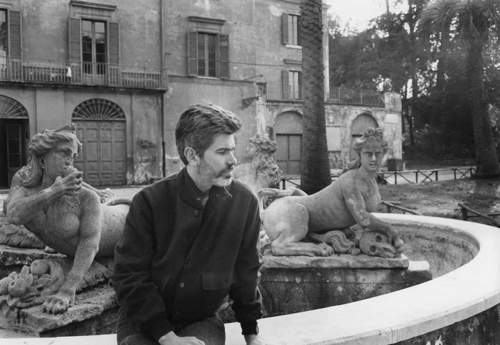 Black-and-white photo of a man sitting on the edge of an old fountain surrounded by weathered sphinx sculptures, with a historic building and palm trees in the background.