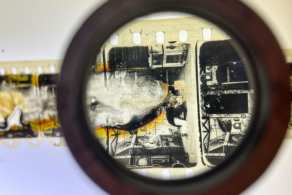 Magnified damaged film strip frame reveals Méliès-style scene with burn marks, machinery imagery, and deteriorating celluloid texture
